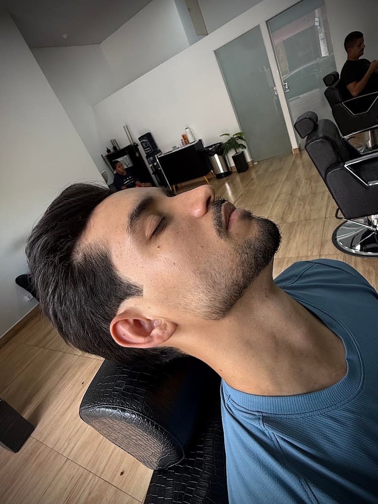 Cliente reclinado na cadeira durante o atendimento, com as demais estações e a entrada da barbearia ao fundo